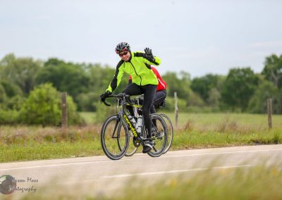 Vibrant cyclist waving on rural bike trail with greenery in background.