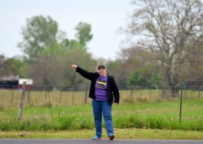 Woman on roadside pointing toward a rural landscape with trees and grassy fields.