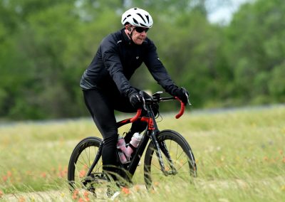 Vibrant cyclist riding a black and red bike across a grassy field with green trees in the background.