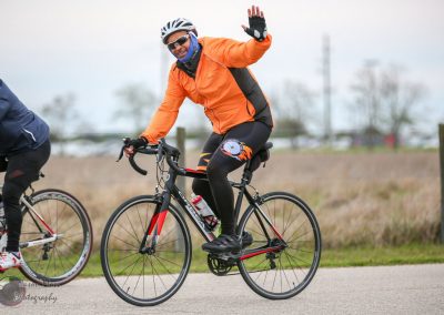 Bright orange cycling jacket, riding a black and red bike, waving during outdoor bike ride.