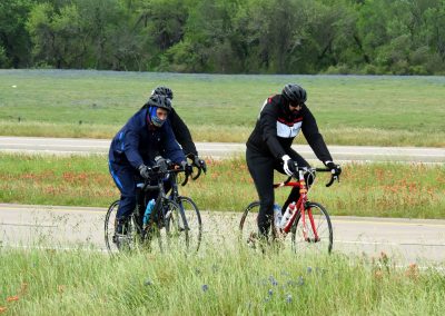 Vibrant cyclists riding on scenic Texas bike trail surrounded by wildflowers and greenery.
