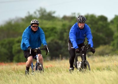 Elderly men cycling outdoors in rain, wearing blue waterproof jackets and helmets.