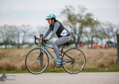 Woman riding a blue road bike during a cycling event, wearing helmet and athletic gear.