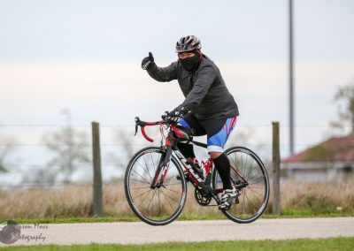 Bright cyclist wearing black gear and a mask, riding a road bike outdoors for Bluebonnet Express.