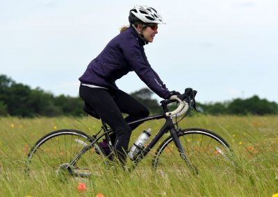 Vegan cyclist riding a black road bike through a grassy field with wildflowers, wearing a helmet and athletic gear.
