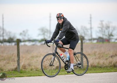 Bright male cyclist riding a road bike on a paved trail in outdoor nature setting, wearing helmet and athletic gear.