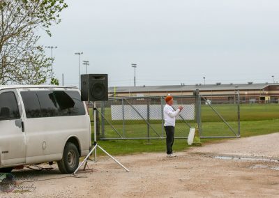 Outdoor event setup with sound system at Bluebonnet Express, featuring a person speaking near a van and large speaker.