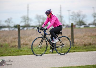 Ally bike rider wearing pink jacket, cycling outdoors on a road with green grass and open landscape.