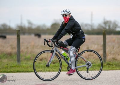 Woman cycling on a Specialized road bike in outdoor setting with grassy fields and a road, promoting Bluebonnet Express bike services.