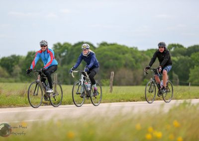 Brightly dressed cyclists riding on a rural bike trail amid lush greenery and open skies, promoting Bluebonnet Express biking adventures.
