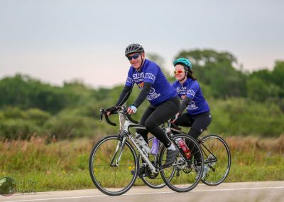Bright blue cycling jerseys with "Sloth" logo on two cyclists riding on scenic green trail during daytime.