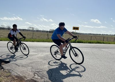 Vibrant cyclists riding on a rural road with a clear blue sky and scenic open fields in the background.