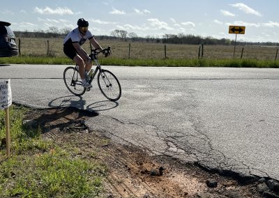 Bike cyclist riding on rural road with cracked pavement and open fields, promoting Bluebonnet Express biking tours.