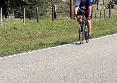 Cyclist riding a bike on a scenic rural road with greenery and trees in the background.