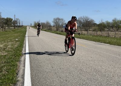Bright women riding bikes on long open road under blue sky near Texas countryside.