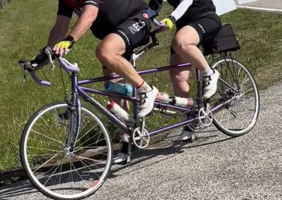 Two cyclists riding a tandem bicycle during a sunny day, wearing matching NEOS jerseys and helmets.