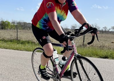 Bright woman riding a pink road bike on a sunny day, wearing a colorful tie-dye jersey and safety gear.