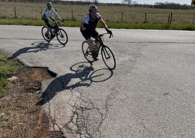 Bicycle riders on a road with a cracked asphalt surface, rural landscape, and blue sky.