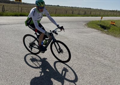 Velo cyclist riding on open country road during sunny day, blue sky, and landscape in the background.