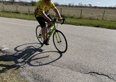 Bright yellow cyclist jersey riding a road bike on open highway with field scenery, sunny weather, and blue sky.