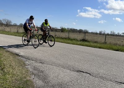 Cyclists riding along a rural road, enjoying a sunny day with open fields and blue sky.