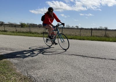 1. Female cyclist riding on scenic country road with blue sky and open fields in the background.
