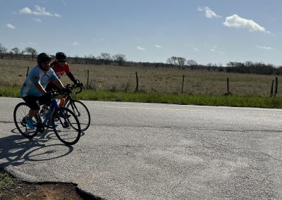 Biking on open road with two cyclists through rural landscape under blue sky and scattered clouds.