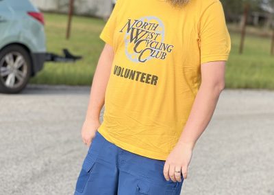 Man wearing a yellow North West Cycling Club volunteer T-shirt and blue shorts outdoors.