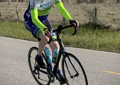 Cyclist riding a blue bike on a rural road with a "Bluebonnet Express" themed jersey, promoting outdoor cycling and endurance.