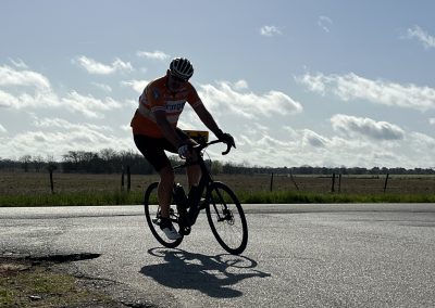 Bright cyclist riding a bike on scenic rural road with blue sky and clouds in the background.