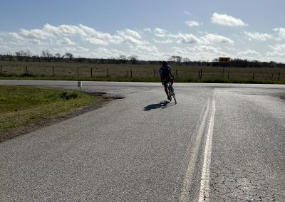 Cyclist riding on a rural road with open fields and blue sky in the background.