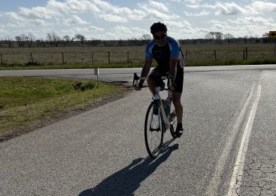 Cyclist riding a road bike on a scenic rural route with blue skies and open fields at Bluebonnet Express.