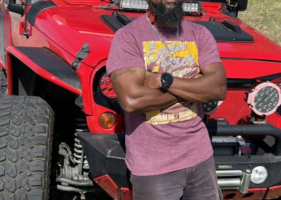 Red off-road vehicle with powerful lights and rugged tires, featuring a man with sunglasses and a cap standing beside it.