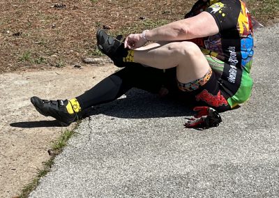 Worn out cyclist sitting on the ground after a ride, wearing colorful cycling gear and helmet, outdoors on a sunny day.