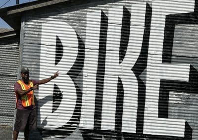 Large "BIKE" sign painted on corrugated metal wall with man pointing at it, promoting cycling awareness.