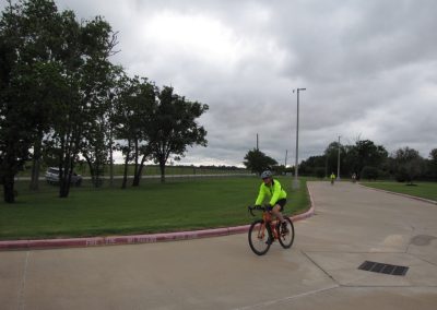 Bicycle rider in neon jacket on trail at Bluebonnet Express, outdoor cycling path with trees and cloudy sky.