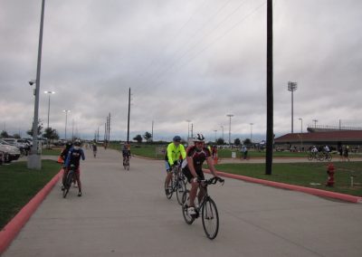 Bright cyclists riding on a paved trail at Bluebonnet Express event, outdoors, with stadium lights and cloudy sky in the background.