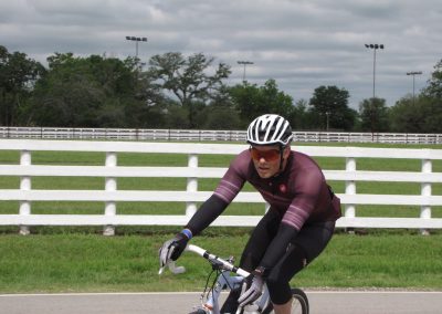 Vegan cyclist riding on a paved path with white fence and trees, training for Bluebonnet Express race.