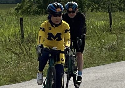 Youth cyclist wearing a University of Michigan jersey riding on a rural road with an adult male cyclist behind.