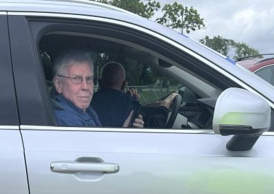 Older man with glasses sits in car, looking at camera while driver holds phone.