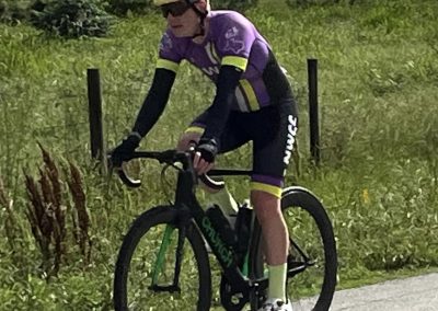 High-performance cyclist riding a road bike in scenic countryside, wearing a purple jersey, helmet, and sunglasses.