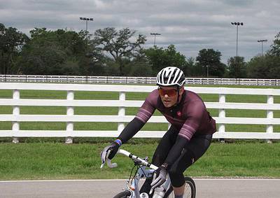 Fast cyclist wearing helmet and sunglasses riding a bike on a trail with a White fence and cloudy sky.
