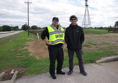 Sheriff and civilian standing together outdoors, rural road, utility poles, and windmill in background.