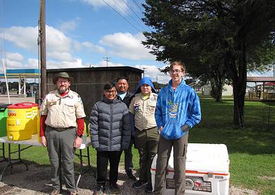 Young girls and boys with Boy Scout uniforms smiling outdoors in front of water coolers and trees, community outdoor event.