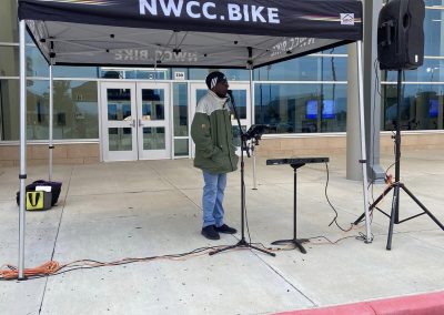 A person speaking at a NWCC Bike event under a black NWCC.BIKE canopy tent outdoors.