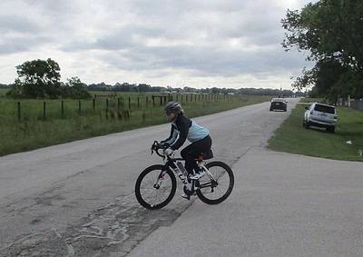 Cyclist riding on rural road with cars parked nearby, scenic Texas countryside, overcast sky, outdoors.
