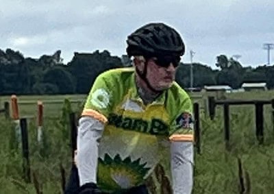 Bike rider in green cycling jersey on rural road with cloudy sky.