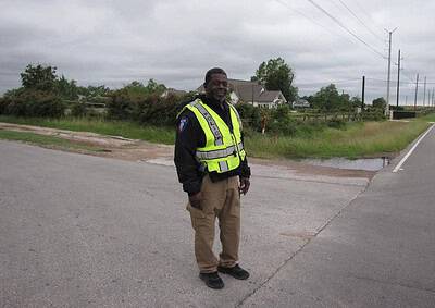 Police officer standing on roadside during daytime, wearing reflective vest, patrol uniform, overcast sky.