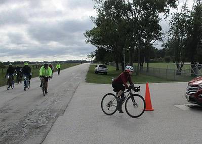 Cyclists riding on a scenic outdoor trail near trees and parked vehicles during daytime.