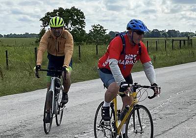 Brightly colored cyclists riding on rural asphalt road with green fields and cloudy sky, promoting Bluebonnet Express cycling event.